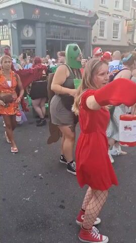 This video shows a street scene with a group of people in costumes. In the foreground, a woman in a red dress and fishnet stockings is dancing. She has blonde hair and is wearing red shoes. Behind her, a person in a green costume with a large headpiece is visible. To the left, a woman in an orange dress with red wings is looking at her phone. Other people are standing around, some of whom are also in costume. The background shows buildings with shops and streetlights. The atmosphere appears festive, possibly during a parade or event. The pavement is wet in places.