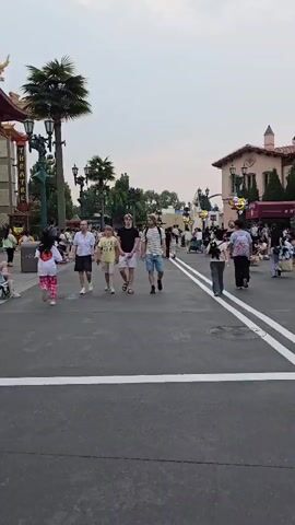 The media file depicts a bustling street scene, likely within a theme park or entertainment district, given the architectural style and the presence of many people. The setting features buildings with a Mediterranean or Spanish colonial influence, adorned with decorative lamps and awnings. Palm trees and other greenery are visible, suggesting a warm or temperate climate. The time of day appears to be late afternoon or early evening, with a bright, overcast sky.

The primary subjects are a diverse group of people walking along a paved pathway marked with white lines. Several individuals are captured in the foreground, including a woman in a white shirt and dark shorts, followed by a group of young men and boys. They are casually dressed, wearing t-shirts, shorts, and sneakers, indicating a leisurely atmosphere. Further in the background, more people are seen walking, some pushing strollers, and others gathered near shops or attractions.

The activity taking place is the movement of pedestrians through an area that seems to be a thoroughfare or plaza. People are casually strolling, suggesting they are enjoying their time and exploring the location. There are no overt signs of a specific event, but the density of people implies a popular attraction or a regular gathering place.

Notable details include a trash receptacle on the left side of the frame and what appears to be a manhole cover with embossed text in the lower right quadrant, which is partially obscured but might indicate a specific location or utility. The overall emotion conveyed is one of a pleasant, active, and public gathering. The scene appears to be taking place in Tongzhou District, China, as provided.