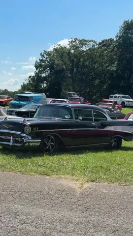 The video shows a sunny outdoor scene, likely a car show or gathering, with a focus on classic cars. Several vintage automobiles, predominantly in shades of red and black, are parked on a grassy field. In the foreground, a black car with chrome accents and custom wheels is prominently featured. Behind it, a row of red cars, including what appears to be a pink Cadillac, are visible. A white sign with red lettering that reads "RICK ROSS" and "PERSONAL VEHICLE ROWS" with directional arrows is placed near the red cars. The background consists of lush green trees and a bright blue sky with scattered clouds. The camera pans slowly across the scene, offering glimpses of various classic car models.