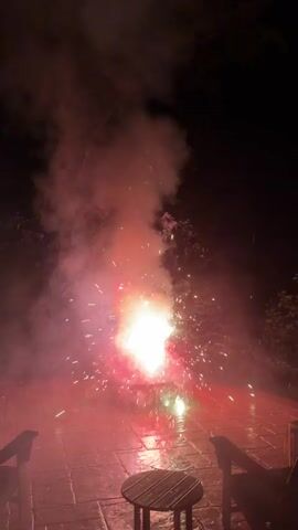 This is a video of fireworks being set off at night on a paved patio. The fireworks create a bright burst of light and sparks, with red, green, and white colors visible. Smoke billows up from the fireworks, obscuring the sky. In the foreground, there are two dark-colored chair backs visible on either side of the frame, and a small, round wooden table with a slatted top is centered in the lower part of the frame. The patio is made of large, square tiles. Trees are visible in the background against the dark night sky.