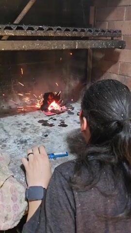 The video shows a person trying to light charcoal in an indoor brick fireplace. The camera focuses on the fireplace, where a small pile of charcoal sits on pieces of red cardboard. The charcoal is starting to burn, with visible flames and glowing embers. Sparks are flying from the fire. The person is seen holding a lighter, but they are not actively using it at this moment. The person has long brown hair pulled back into a ponytail and is wearing a grey shirt. A watch is visible on their left wrist. The floor in front of the fireplace is speckled with dark stains. A metal grill sits above the fire. Music is heard in the background. The person states the process of starting the fire is proving to be difficult.