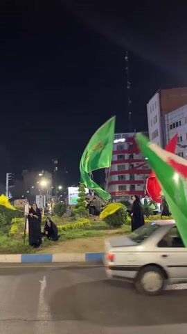 A nighttime street scene features several individuals, some in black abayas, holding and waving flags. The flags are colorful, with one prominently displaying red, white, and green horizontal stripes, and another a bright green. Yellow flags are also visible. A white car drives across the foreground, its headlights illuminated. The background includes buildings and streetlights. The overall atmosphere suggests a public gathering or event.