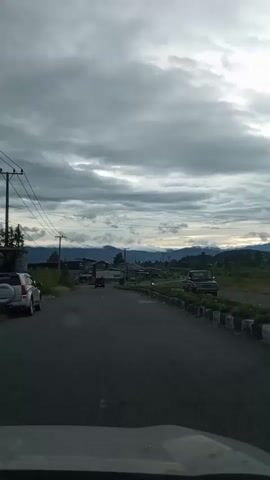 The video shows a road with several vehicles. On the left side of the road, there is a white truck driving away from the camera. In the distance, there are some buildings and hills. On the right side of the road, there is a black pickup truck parked facing the camera. There are also some motorcycles driving on the road. The sky is cloudy and appears to be overcast.