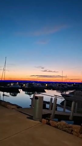 The video shows a marina at dusk. The sky transitions from a deep blue at the top to orange and pink near the horizon. Numerous sailboats and motorboats are docked in the water, their lights illuminated, reflecting on the calm surface. A dark pole is prominently in the foreground on the right. To the left, a trash can and part of a walkway with a railing are visible. The walkway is bordered by large rocks. A person wearing a green shirt and denim shorts walks away from the camera along the walkway. The video is a still shot that does not depict any actions or interactions.