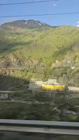This media file captures a scenic view of a hydroelectric dam nestled within a mountainous region. The dam itself is a substantial concrete structure with a striking bright yellow building on top, possibly housing operational equipment. Water is visible flowing from the dam, creating a reservoir.

The surrounding setting is characterized by lush green, rolling mountains, their slopes dotted with vegetation and rocky outcrops. The sky above is a clear blue with scattered white clouds, suggesting fair weather. The sunlight casts shadows on the mountains, indicating it's likely daytime, possibly mid-morning or afternoon.

The perspective suggests the recording was made from a moving vehicle, likely a train, as indicated by the blurred foreground and the guardrail visible at the bottom of the frames. This motion adds a sense of travel and journey to the visual experience. The location is identified as Nizhnii Zaramag, Russia, known for its mountainous terrain and hydroelectric power generation.

There are no visible people in the frames, and the focus is on the natural landscape and the industrial structure of the dam. The overall mood is one of impressive scale and natural beauty, with the man-made dam harmoniously integrated into the rugged environment. There is no visible text within the media file.