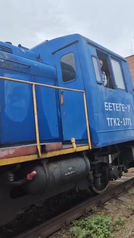 This media file shows a close-up, low-angle view of a blue diesel locomotive and adjacent green freight cars on a railway track. The locomotive, labeled "56-0008" on its side, has its engine compartment partially open, revealing the engine and some red tool holders. A person is visible in the locomotive's cab, looking out. The freight cars appear to be grain hoppers, one of which has the Cyrillic word "ЗЕРНО" (grain) visible. The scene is outdoors, under an overcast sky, suggesting a cloudy day. The setting appears to be an industrial area or railyard in Petropavl, Kazakhstan, with a tall white building and trees in the background. The overall impression is one of industrial activity, possibly related to maintenance or operation of the train.