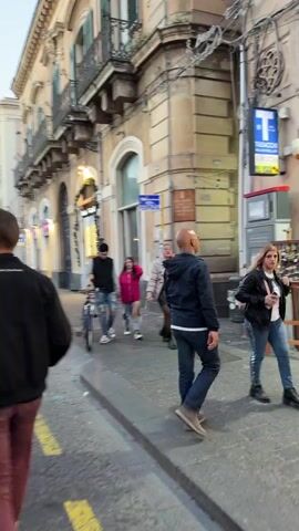 The video shows a street scene with pedestrians, including a man walking a bicycle, people with a baby stroller, and other individuals walking in various directions. The street features buildings with architectural details, a bus stop sign, and trees along the sidewalk. Some people are sitting at outdoor cafes. The overall setting appears to be a European city.