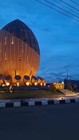 The video shows a low-angle shot of a large, egg-shaped building at dusk. The building is illuminated with warm yellow lights, highlighting its textured surface and a perforated upper section. Surrounding the base of the building are several smaller, drum-like structures, also lit from below. In the foreground, large yellow letters spell out "SIGLI" on a tiered platform, with "Bank Aceh" visible in smaller letters in front of the building. The sky is a deep blue, indicative of twilight. The camera pans from left to right, revealing the entire structure and its surroundings.