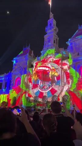This video captures a dazzling nighttime light show projected onto the facade of the Ho Chi Minh City Hall in Vietnam. The architecture of the grand building serves as a canvas for a vibrant and dynamic projection, featuring abstract patterns and a central, stylized image that resembles a mask or a mythical creature with a fierce expression. The projected imagery utilizes a bold color palette of red, green, blue, and yellow, creating a lively and energetic display.

A crowd of people is gathered in the foreground, silhouetted against the illuminated building. Many are holding up their phones to capture the spectacle, indicating the event's popularity and the audience's engagement. The time of day is clearly night, with the darkness of the sky providing a stark contrast to the bright, colorful lights. The scene evokes a sense of wonder and excitement, as the crowd experiences a shared moment of visual entertainment.

The national flag of Vietnam is visible at the very top of the building's clock tower, adding a patriotic element to the scene. The overall atmosphere is festive and celebratory, likely part of a public event or festival. The projection is intricate and detailed, showcasing advanced lighting and mapping technology. The building itself is a prominent landmark, recognized by its classical French colonial architecture, including the clock tower and ornate facade details. The light show transforms this historical structure into a modern spectacle.