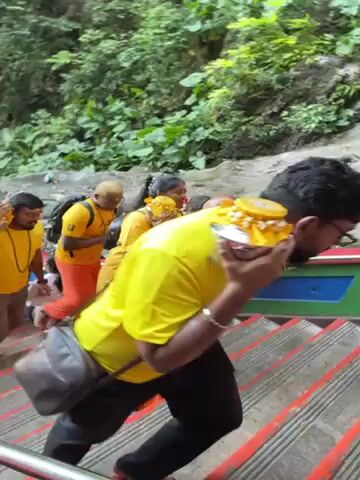 A group of people is walking up a steep, colorful staircase in an outdoor setting. The individuals are wearing yellow shirts and either orange or grey pants. Most carry a small metal pot or container on their heads, adorned with yellow decorations. Lush green vegetation is visible along the sides of the staircase, indicating a natural, possibly tropical environment. The group is chanting "Vel Vel," suggesting a religious or ceremonial procession. The staircase appears well-maintained and painted with vibrant colors, contrasting with the surrounding natural setting.