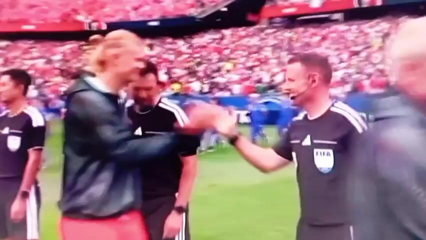 The video shows a group of soccer players and officials on a soccer field with a crowd in the background. In the first frame, a referee is shaking hands with a player who has long blond hair. In the subsequent frames, a team of players in white uniforms with blue trim are lined up, some of whom are shaking hands with each other and with officials. The stadium is filled with spectators wearing red. The environment appears to be a professional soccer match.