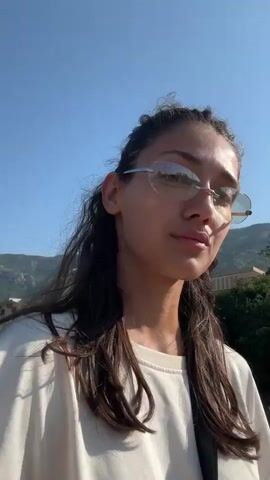 A close-up, medium shot shows a young woman from the chest up, looking up and to the right, then turning her head to look down and to the left towards the camera. She has long, dark, wavy hair pulled back loosely, and is wearing large, rimless sunglasses with a silver frame. She is also wearing a white t-shirt. The background is a bright blue sky with some green trees and buildings on the right. The lighting suggests it is daytime.