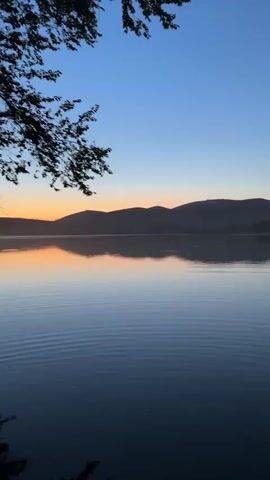 This is a vertical video of a serene lake at dawn. The sky transitions from a deep blue at the top to a soft orange and pink glow near the horizon, indicating sunrise. Silhouetted hills form the backdrop, with several antennas visible on their crests. The calm lake mirrors the sky and the distant landscape, creating a symmetrical and peaceful scene. The water has gentle ripples, suggesting a light breeze. On the left side of the frame, a dark tree branch with leaves hangs over the water, adding a natural frame to the composition. The overall mood is tranquil and reflective. No people or specific objects are clearly visible other than the natural elements and antennas. The video is undated and has no discernible text. The location is identified as Milyanfan, Kyrgyzstan.