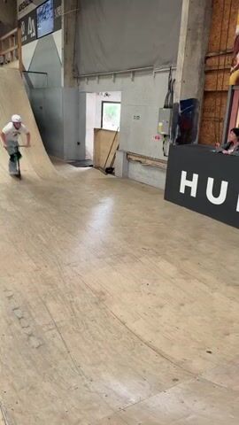 A young person wearing a white helmet, t-shirt, and light-colored pants rides a scooter on a wooden ramp in an indoor skatepark. The person rides up one side of the ramp, performs a trick in the air, and lands back on the ramp, continuing to ride. The skatepark has various ramps and obstacles, and there are people in the background watching. A sign with the word "HUFFY" is visible.