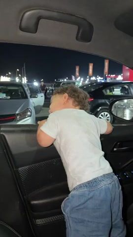 A child with curly brown hair wearing a white t-shirt and blue jeans leans out of a car window at night. The child's back is to the camera, and they are looking to the left. In the background, there are several cars parked in a lot with lights and buildings visible. The interior of the car, including the door panel and handle, is also visible.