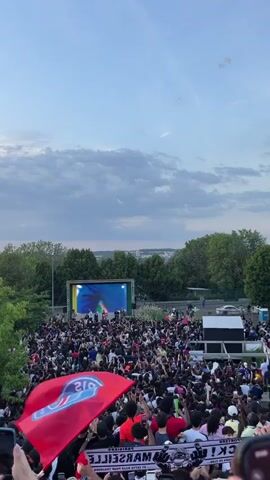 The video shows a large crowd gathered outside watching a soccer game on a big screen. Many are waving a red flag with a blue logo, presumably the team they are supporting. There are also what look like streamers being thrown up into the air. The people are dense and seem to be excited or celebratory. Trees border the area, and a cloudy sky is overhead.
