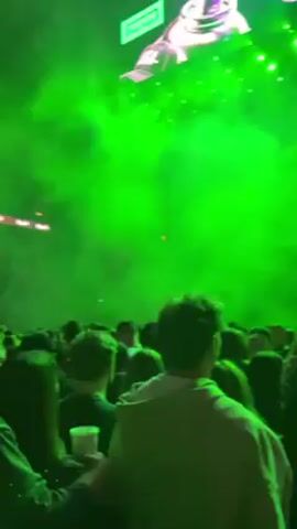 A vertical video shows a crowd of people at a large concert in a stadium. The view is from within the crowd, looking toward the stage, which is obscured by green lighting effects, including smoke. The crowd appears to be mostly adults, dressed casually. Some are holding drinks. The stadium is full, with people seated in the upper levels. The roof of the stadium is visible, with a geometric metal framework. The overall lighting is dominated by the bright green lights from the stage.