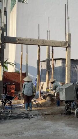 This video shows a construction site. Two individuals, both wearing conical hats and long-sleeved shirts, are working. One individual is standing and appears to be observing, while the other is kneeling and actively working with what appears to be cement or sand. There are construction materials visible, including bricks, rebar, and wooden beams used for support. A cement mixer is also present. The scene suggests ongoing construction work.