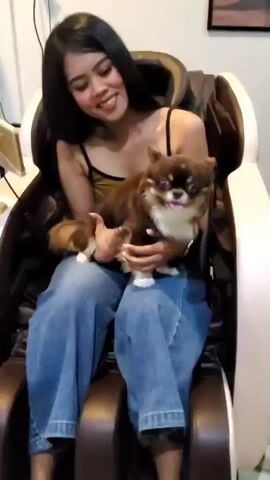 A young woman sits in a massage chair holding a small, long-haired dog on her lap. She is wearing a black tank top with spaghetti straps and blue jeans. The dog is brown and white with its tongue sticking out. The woman is smiling and looking at the dog. The massage chair is brown and beige. There is a guitar and a tablet on a stand behind the woman. The room appears to be a living room or a salon.