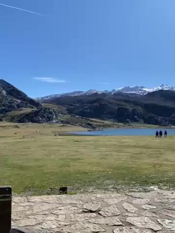 The video shows a serene landscape. The foreground is a paved stone surface, possibly a pathway or viewing area. Beyond that is a wide expanse of grassy field, leading to a calm lake. In the lake's background are dark, rugged mountains, some with visible patches of snow on their peaks. Four figures are seen walking near the lake's edge, appearing to be people enjoying the scenery. The sky above is clear and blue with the contrail of an airplane visible across the top.