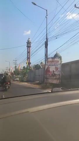 The video shows a street view from inside a vehicle. A tall mosque minaret dominates the background, with a domed mosque structure visible to its right. The street is lined with utility poles and wires. Several flags, including Indonesian flags, are displayed along the roadside. There are vehicles on the road, including motorcycles and cars. One car has a banner on its roof. The scene appears to be in a sunny, outdoor environment.
