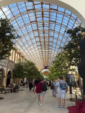 The video shows a well-lit, indoor walkway with a high, arched glass ceiling. The ceiling structure is a network of golden metal beams. Sunlight filters through the glass, illuminating the walkway. On either side of the walkway are rows of tall, leafy artificial trees, creating a canopy effect.

Several people are walking in both directions along the walkway. Most of them are seen from behind, dressed in casual summer clothing. Some individuals are carrying backpacks. In the foreground, two men are walking away from the camera. One is wearing a white t-shirt, shorts with a floral pattern, and a backpack with blue accents. The other man is wearing a maroon t-shirt, dark shorts, and a black backpack. Further ahead, more people are visible, including a woman pushing a stroller.

On the right side of the walkway, a vertical sign with illuminated numbers, "13," is mounted on a pole. On the left side, there are buildings with arched entrances and some decorative elements.

Suspended from the ceiling, in the middle of the walkway and high above the people, is a large, purple, balloon-like inflatable object shaped like a mythical creature or a cartoon character. It appears to be a focal point or decoration within the space. The overall atmosphere appears to be that of a bustling indoor public space, possibly a shopping mall or a conservatory.