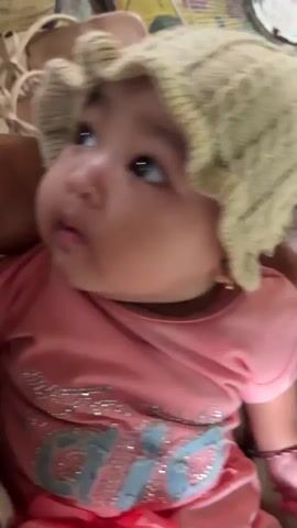 A baby wearing a knitted hat and a pink shirt is being held. The baby is looking around. The background is blurred but appears to be a room with a fan and some patterned fabric.