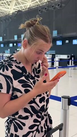 A young woman with blonde hair pulled into a bun, wearing a black and white patterned dress, stands at an airport check-in counter. She is holding an orange smartphone in her right hand and using her left hand to rub her eye. Her face shows a look of distress or discomfort. In the background, there are airport check-in desks and barriers.