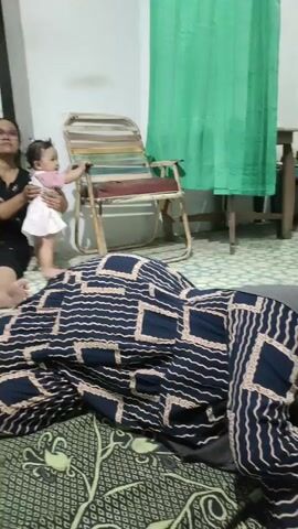 A woman wearing glasses and a black top is sitting on the floor, holding a baby. The baby is wearing a pink and white dress and is reaching for a chair. The chair is a folding chair with colorful stripes. A patterned blanket is in the foreground. Another person's legs are visible to the left. The room appears to be indoors, with a tiled floor and a white wall. A green curtain is visible in the background.