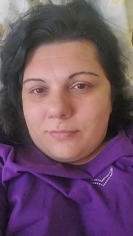 This is a first-person point-of-view video. The camera is pointed at the face of a woman lying down. She has dark hair and is wearing a purple hoodie. The background appears to be a bed.