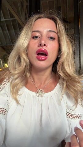 A blonde-haired woman wearing a white blouse and a silver necklace is talking directly to the camera, with her hands raised near her face. She purses her lips slightly at one point. The background is blurred, but appears to be inside a building with a high, beamed ceiling and a dark-framed window or door.