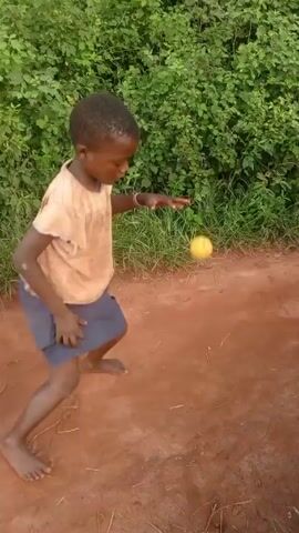 Here is a brief description of the video:

The video shows several children playing outside on a reddish-brown dirt surface. One child kicks a yellow fruit, possibly a mango or similar, while another child holds a similar fruit. A third child is sitting on the ground, and an adult passes by in the background. The backdrop is lush green vegetation. The children are barefoot and dressed in casual clothes.