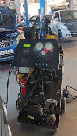 This video shows a car repair shop with a machine in the foreground. The machine has a digital display, two gauges, a keypad, and various hoses and components. It appears to be an air conditioning service machine for vehicles. In the background, there are several cars, including a blue Honda with its hood open and a silver Smart car. A person wearing a dark shirt and jeans is visible in the background, standing near a counter and a laptop. The environment is a typical garage setting with fluorescent lighting and concrete flooring. The machine is on wheels, suggesting it is portable within the shop.