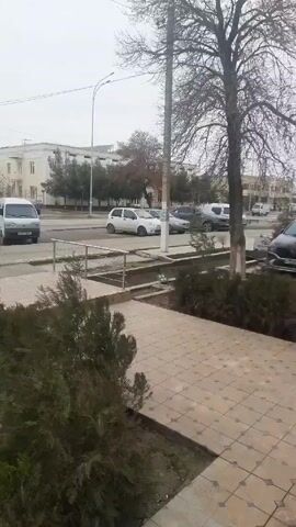 The video shows a street scene in what appears to be a small town. The weather is overcast. There are several cars visible on the road, including a white van, a gray sedan, and a black sedan. A person is riding a bicycle on the sidewalk. There are buildings on either side of the road, and trees and shrubs planted in the median strip. The video appears to have been taken from an elevated position.