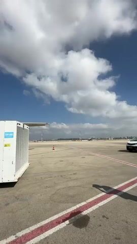The video shows an airport tarmac scene. In the foreground, a large white air conditioning unit is visible on the left. An airplane, identified by the text "ISRAIR" on its fuselage and an orange and blue tailfin, is parked. Passengers are boarding the aircraft via a mobile staircase on the left. A baggage tug is positioned near the airplane's nose. A person wearing a high-visibility vest stands near the staircase. The sky is blue with scattered white clouds. The ground is asphalt with red and white markings.