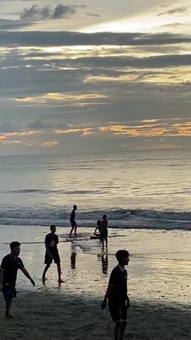The video shows a beach scene at dusk. Several people are visible on the wet sand and in the shallow water. In the foreground, four young men are standing and walking. One is in a black t-shirt and shorts, walking towards the left. Another in a black t-shirt and shorts is facing right, with his right arm raised as if to throw something. A third person in a dark shirt and shorts is bent over. A fourth person, also in dark clothing, is in the background to the left. Further out in the water, three silhouetted figures are standing. One is in the shallow waves, and two are further out, one appearing to be holding a surfboard. The sky above is filled with dramatic clouds, with streaks of orange and yellow light suggesting the setting sun. The ocean is calm with gentle waves lapping the shore. The overall atmosphere is serene and suggests a recreational activity taking place at the end of the day.