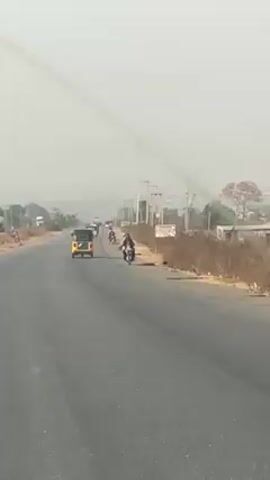 The video shows a highway during the day under a hazy sky. The road is asphalt and has some visible wear. A yellow auto-rickshaw (tuk-tuk) travels in the same direction as the camera, while a motorcyclist goes in the opposite direction. There are power lines running along the side of the road. The land adjacent to the road is mostly dry and barren, with some scattered trees and shrubs. A person is seen walking alongside the road carrying something on their head. Other vehicles can be seen further down the road.