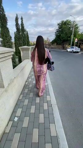 A person in pink pajamas walks away from the camera on a paved sidewalk next to a street. The person has long dark hair and carries a black bag over their left shoulder. A white car is parked on the street in the background. The sky is cloudy with blue patches.