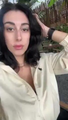 A woman with dark hair and light skin is recording a selfie video outdoors, likely on a patio or in a courtyard. She is wearing a light-colored jacket and appears to be sitting. The background includes plants, chairs, and what looks like a corrugated metal roof or awning. She runs her hand through her hair several times during the video. Music can be heard in the background.