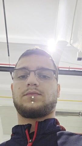 A close-up selfie-style video depicts a young man with short brown hair, wearing glasses, a navy and red jacket, and a beard. He stands in a large, brightly lit industrial space. Behind him are several pieces of large, white machinery with screens and controls, suggesting a manufacturing or production facility. There are also rolling carts and other industrial equipment visible. The camera moves slightly as the man holds it, showing more of the environment and then refocusing on his face. The lighting is predominantly bright and overhead, creating some glare in the shots.