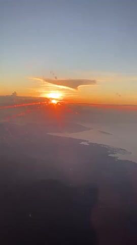 The video shows a view from an airplane window at sunrise or sunset. The sun is partially obscured by clouds, creating a vibrant orange and red glow. Below the clouds, land and water are visible, with the land appearing darker and the water reflecting the light. A finger briefly appears in the bottom left corner of the frame. The sky above is clear blue, transitioning to a lighter hue near the horizon. The window appears to have some minor smudges or imperfections.