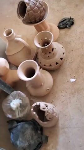 The video shows a collection of unglazed pottery vessels lying on a dusty, light-colored floor. The pieces vary in shape and size, including vases with wide flaring openings, one with a handle, and one with decorative cutouts. Some pieces are wrapped in plastic. A metal ladder and other objects are visible in the background, suggesting a workshop or storage area. The video appears to be shot at a low angle, panning out to reveal the full collection of pottery.