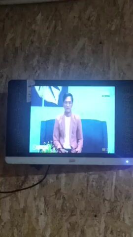 A television screen displays a man wearing a pink blazer over a white t-shirt and black pants. He is seated on a dark couch. A small plant is visible on a table to his left. The background is a light blue. The timestamp on the screen reads 08:36. The television is mounted on a wood-paneled wall.