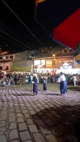 At night, three people in traditional attire dance on a cobblestone plaza surrounded by a crowd. The dancers wear dark-colored robes, wide-brimmed hats, and long skirts. One holds a staff. The plaza is lit by streetlights and colorful spotlights. Buildings with lights and windows surround the plaza. The edge of a large, colorful umbrella is visible in the foreground.