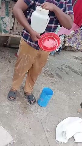 Here is a detailed description of the video:

The video shows a young boy standing in a cluttered outdoor area. He is holding a white plastic container with a red plastic base, likely a chicken feeder, and smiling at the camera. He is wearing a plaid shirt, tan pants, and sandals. A blue plastic container is on the ground beside him. In the background, a chicken pecks at the ground. Laundry hangs on a line in the background. The ground appears to be concrete.