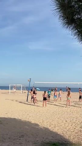 The video shows a beach scene with a group of people playing volleyball. The sky is clear and blue with a few clouds. The sand is light brown. There is a volleyball net set up in the middle of the frame. Several people are actively playing volleyball, with others observing or standing nearby. In the background, there is a body of water, presumably the ocean, and a beach with other people. There are also some trees or foliage visible in the foreground on the right side of the frame. The video captures a moment of recreation and activity on a sunny day at the beach.