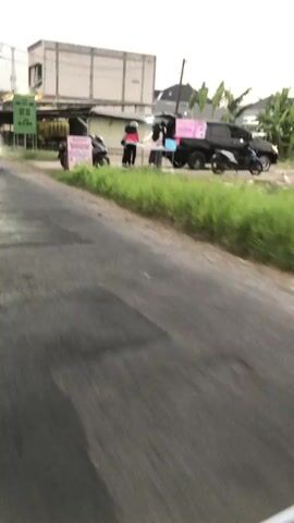 The video captures a street scene in Boro, Indonesia, likely during the late afternoon or early evening due to the soft, diffused lighting. The weather appears overcast. The scene is set along a paved road with a grassy verge.

The primary subjects are several people and motorcycles. On the left side of the road, a person rides a motorcycle, moving away from the camera. In the middle ground, a small roadside stall is set up. Two individuals are visible behind a table, possibly attending to customers or preparing items. A black pickup truck, modified to serve as a mobile shop, is parked behind the table, with its rear open. A pink sign is prominently displayed on the truck. A black motorcycle is parked next to the stall. Another person on a motorcycle is seen further down the road, also moving away from the camera. In the foreground, the side mirror of a motorcycle is visible, suggesting the footage is being recorded from a moving motorcycle.

The activity taking place appears to be a small business operation at the roadside, likely selling food or drinks, as indicated by the stall setup and the mobile truck. The general atmosphere seems to be that of daily life and commerce in a semi-urban or rural Indonesian setting.

A visible sign near the stall has text in Indonesian. It reads "DONUT KETAN HITAM" which translates to "Black Sticky Rice Donut". This suggests that donuts made with black sticky rice are being sold. The overall impression is one of a casual, everyday scene with a focus on a small entrepreneurial venture.