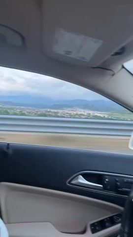 A young woman is visible in the interior of a moving car, on a highway, making kissing faces towards the camera. The car's interior features beige leather and a black dashboard. Outside the window, a landscape with buildings and mountains is visible under a partly cloudy sky. The highway has a metal barrier. There is music playing.