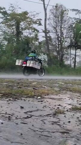 A person on a motorcycle with two large white boxes on the back rides down a wet, dirt road in the rain. The person is wearing a helmet and a green and blue jacket. The road is surrounded by trees and vegetation. There is a utility pole with wires on the right side of the frame. The rain is heavy, creating a mist on the road.