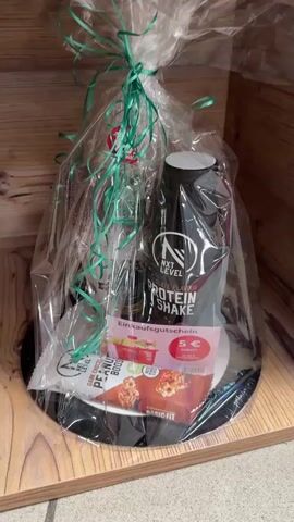 The video shows a close-up of a gift basket wrapped in cellophane and tied with green ribbon. The basket contains a black bottle labeled "NXT LEVEL PROTEIN SHAKE," a small bottle, a snack bar with "DARK CHOCOLATE PEANUT BOOST" and "BASIC FIT" printed on it, and a pink voucher that says "Einkaufsgutschein 5 € rabatt." The basket sits on a black circular base, which is placed on a wooden shelf. The background is a wooden surface. The camera then pans to the right, revealing more gift baskets on wooden shelves in a store-like setting. One basket has a white wrapping with red hearts and a bottle. Another basket, on a lower shelf, is filled with various items and tied with yellow and red ribbons. The top shelf shows a basket with yellow and pink ribbons and a light-colored filling. The video ends with a view of a gift bag on the bottom shelf.